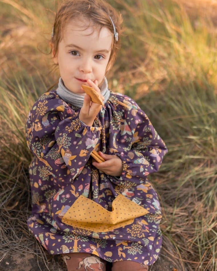 Niña con bata de flores moradas y bolsillo amarillo entre la hierba